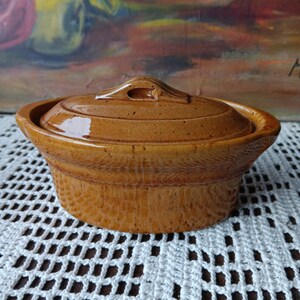 May include: A brown ceramic casserole dish with a lid. The dish has a rounded shape with a textured exterior and a handle on the lid. The dish is sitting on a white crocheted surface.