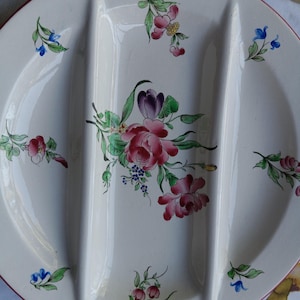 May include: A round white ceramic serving dish with two raised dividers creating three sections. The surface is decorated with hand-painted floral motifs in shades of pink, purple, green, and blue, featuring a thin red rim around the edge.
