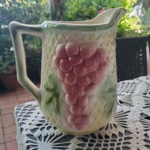 May include: A vintage ceramic pitcher with a grape design. The pitcher is white with a green vine and pink grapes.