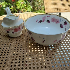 May include: A white porcelain serving bowl and creamer set. The bowl and creamer are decorated with pink floral patterns and gold trim. The set is placed on a woven surface.