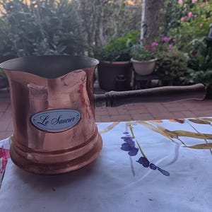 May include: A copper sauce pot with a dark wooden handle and a pouring spout. The exterior features a prominent oval white label with a blue border and the text Le Saucier printed in black script. The interior of the pot is metallic silver.