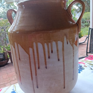 May include: A large, handcrafted ceramic vase with two handles. The vase features a two-tone design, with a rich brown glaze at the top that drips down over a creamy white base. The vase is likely used for decorative purposes.