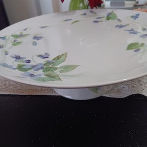 May include: White ceramic cake stand with a floral design featuring blue flowers and green leaves. The stand has a wide, flat top and a short, rounded base. The floral pattern is delicate and adds a touch of elegance to the serving piece.