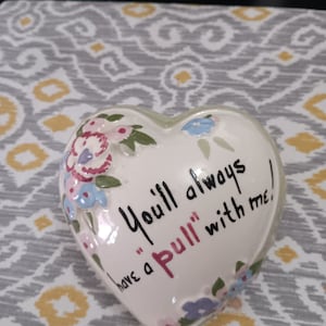 May include: White ceramic heart-shaped trinket with a floral design and the text "You'll always have a "Pull" with me!"