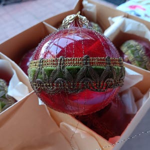 May include: A red glass Christmas ornament with gold filigree and a green velvet band. The ornament is in a cardboard box with other ornaments. The ornament has a gold cap and is round in shape.