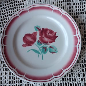 May include: White ceramic plate with a burgundy scalloped rim and a burgundy rose floral design. The rose has green leaves and stems. The plate is on a white crocheted tablecloth.