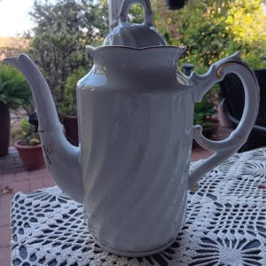May include: A white porcelain teapot with a lid, featuring a spiral design and gold trim. The teapot has a curved spout and a handle with a decorative design. The background is blurred, suggesting an outdoor setting.