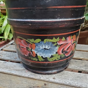 May include: A black painted wooden planter with a red, orange, blue, and green floral design. The planter has a wide rim and a rounded base.
