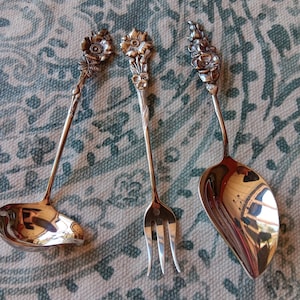 May include: Three pieces of ornate silverware: a ladle, a fork, and a serving spoon. Each piece has a floral design at the handle's end. The silverware is made of a shiny, reflective metal, likely silver or a silver-toned alloy.