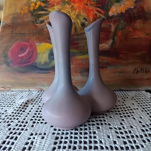 May include: Three slender, pastel-colored vases with a bulbous base and flared top. The vases are a soft pink and blue gradient. They are set against a backdrop of a floral painting and a white crocheted doily.