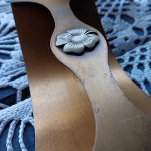 May include: Close-up of two copper-colored metal decorative pieces. Each piece has a curved shape with a floral embellishment in the center. The background is a white lace doily.