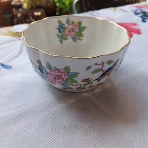 May include: A white porcelain bowl with a scalloped rim and gold trim. The bowl is decorated with pink and blue floral and bird motifs. The bowl is sitting on a white tablecloth with a floral pattern.