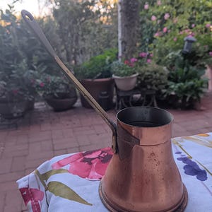 May include: A copper coffee pot with a wide, rounded base and a long, slender metal handle attached to the side. The pot features a cylindrical upper section and a tapered body, showing a metallic finish with visible signs of wear and oxidation.