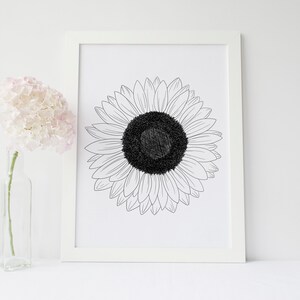 May include: A framed black and white sunflower art print. The sunflower is centered in the frame, with a detailed outline of the petals and a textured black center. A clear glass vase with pink flowers is to the left of the frame.