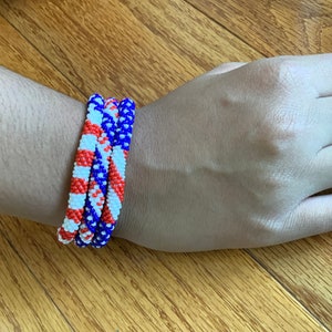 Puede incluir: Tres pulseras de cuentas apiladas en una mu&ntilde;eca. Las pulseras son rojas, blancas y azules, con un patr&oacute;n diagonal.