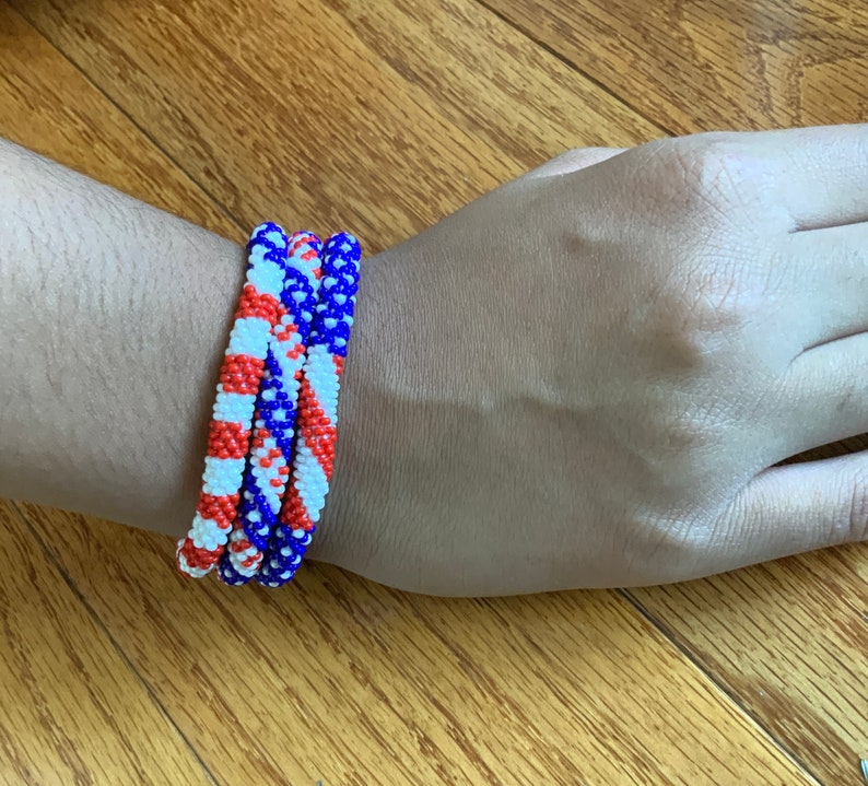 Puede incluir: Tres pulseras de cuentas apiladas en una mu&ntilde;eca. Las pulseras son rojas, blancas y azules, con un patr&oacute;n diagonal.