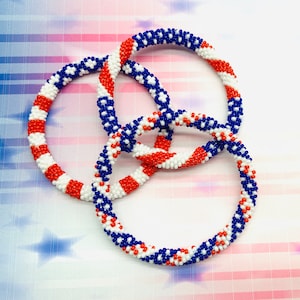 Puede incluir: Tres pulseras de cuentas en colores rojo, blanco y azul. Las pulseras est&aacute;n tejidas juntas en un patr&oacute;n circular.