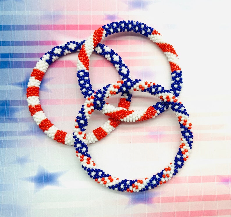 Puede incluir: Tres pulseras de cuentas en colores rojo, blanco y azul. Las pulseras est&aacute;n tejidas juntas en un patr&oacute;n circular.