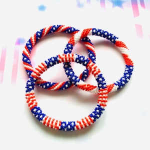 May include: Three beaded bracelets in red, white, and blue, arranged in a circular pattern. The bracelets are made of small beads, with a spiral design. The colours evoke a patriotic theme.
