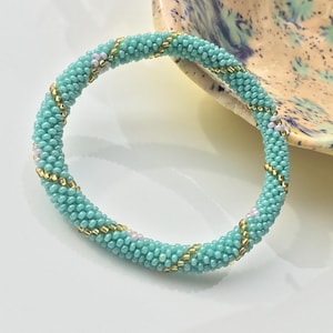 Könnte beinhalten: Ein türkisfarbenes Armband mit goldenen und weißen Akzenten. Das Armband besteht aus kleinen Perlen und hat ein Spiraldesign.