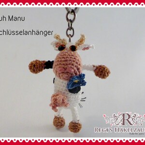 May include: A handmade crochet cow keychain with a metal ring. The cow is white, pink, and brown, with a blue flower and black accents. The text "Kuh Manu Schlüsselanhänger" is visible.