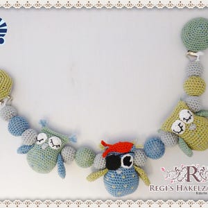 May include: Crocheted baby toy featuring three owl figures in blue, green, and yellow, with gray and blue spheres. One owl wears an eye patch and a red hat. The toy has a clip and the text "REGI'S HÄKELZAUBER".