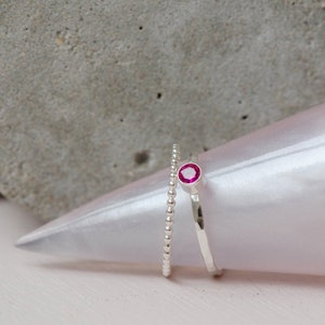 May include: Two silver rings, one with a beaded design and the other with a hammered finish. The beaded ring has a small, bright pink gemstone.