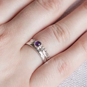 May include: A set of three silver rings. The top ring features a small, round, purple gemstone. The middle ring has a hammered texture, and the bottom ring has a beaded design. The rings are stacked on a finger.