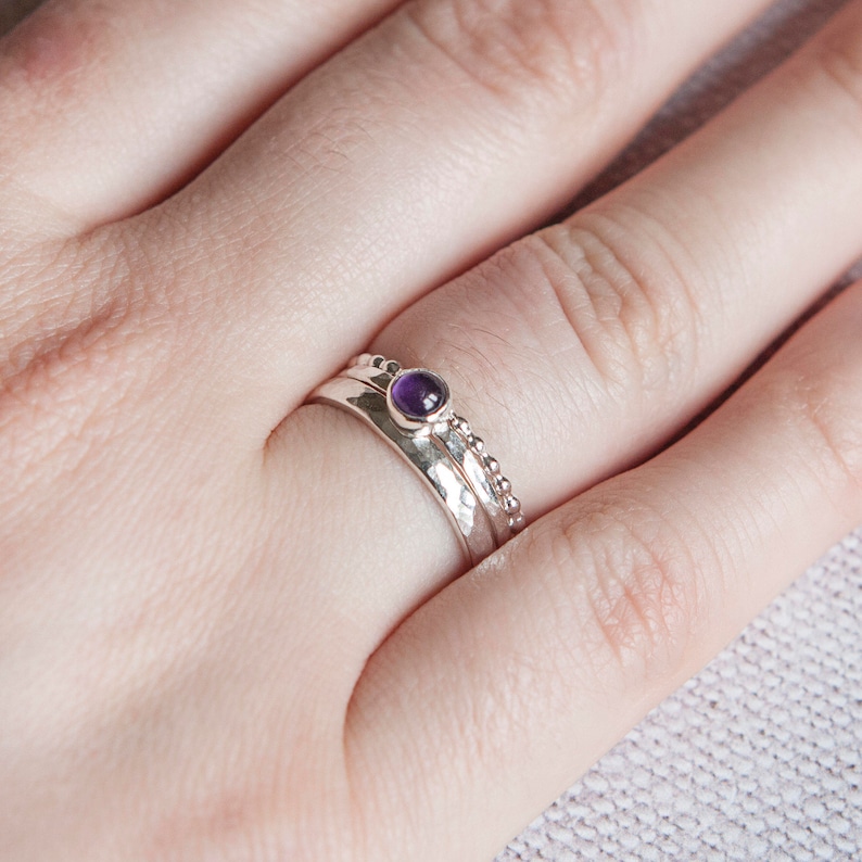 May include: A set of three silver rings. The top ring features a small, round, purple gemstone. The middle ring has a hammered texture, and the bottom ring has a beaded design. The rings are stacked on a finger.