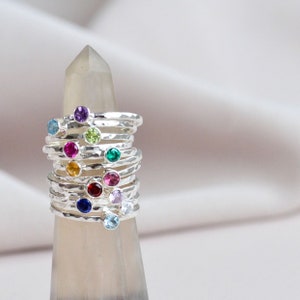 May include: A stack of silver rings with colourful gemstones. Each ring has a different coloured gemstone, including blue, green, red, purple, and yellow.