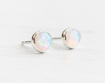 May include: A pair of stud earrings featuring round, iridescent opal stones set in silver bezels. The opals display a range of colors, including white, pink, and blue, with a subtle shimmer. The earrings are displayed on a white surface.