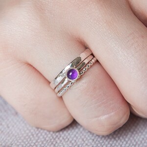 May include: A set of three silver rings. One ring features a hammered texture, another has a beaded design, and the third holds a round, vibrant purple gemstone. The rings are stacked together on a finger.