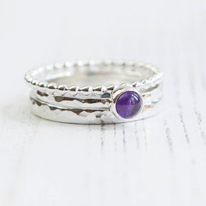 May include: A stack of three silver rings. The top ring has a beaded design, while the middle ring has a hammered texture. The bottom ring has a hammered texture. The middle ring features a round, purple gemstone.