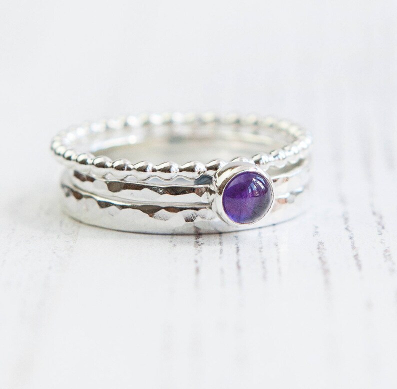 May include: A stack of three silver rings. The top ring has a beaded design, while the middle ring has a hammered texture. The bottom ring has a hammered texture. The middle ring features a round, purple gemstone.