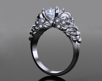 Puede incluir: Un anillo negro con un diamante grande y claro y dos acentos en forma de calavera. El anillo está engastado con diamantes más pequeños alrededor de las calaveras.