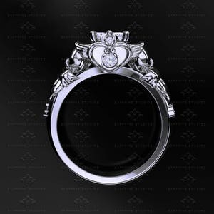 Puede incluir: Un anillo de plata con una piedra central en forma de coraz&oacute;n y dos alas a cada lado. El anillo est&aacute; engastado con un diamante redondo y transparente en el centro del coraz&oacute;n.