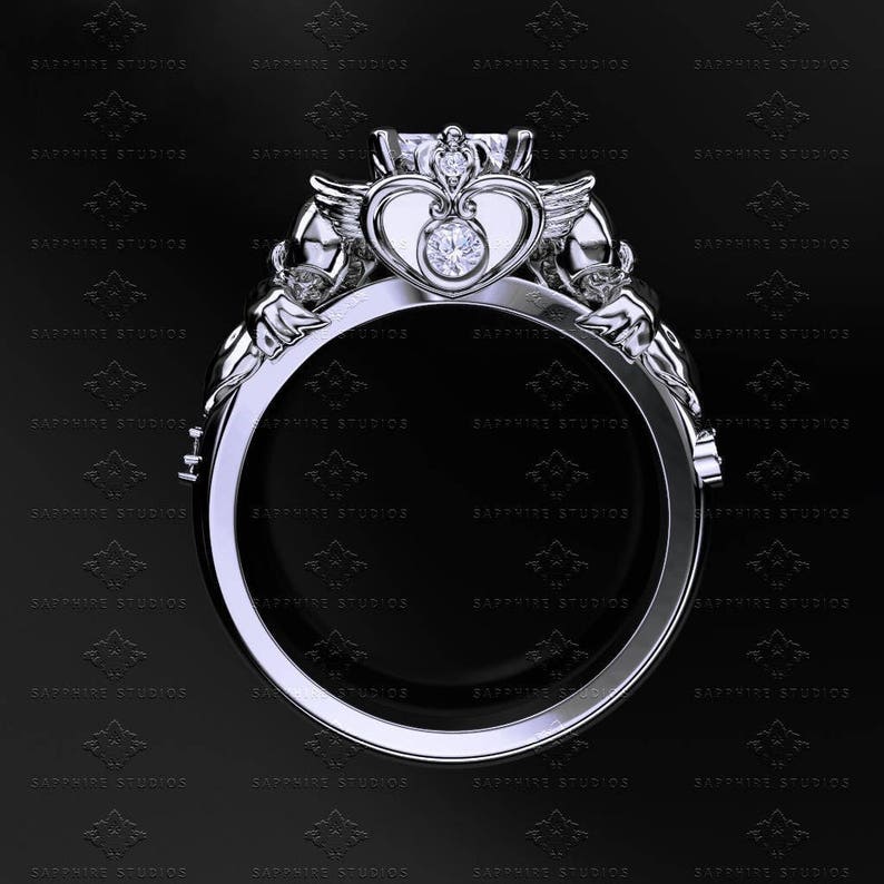 Puede incluir: Un anillo de plata con una piedra central en forma de coraz&oacute;n y dos alas a cada lado. El anillo est&aacute; engastado con un diamante redondo y transparente en el centro del coraz&oacute;n.