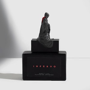 以下が含まれることがあります： 黒い香水瓶。黒と赤のラベルには「INFERNO 50ml/1.7oz. SAPPHIRE STUDIOS」と書いてあります。瓶の上には黒い人形が乗っています。