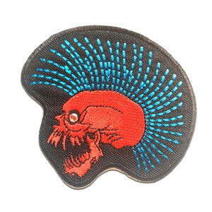 Op de afbeelding: Rode en blauwe geborduurde patch van een schedel met een mohawk. De schedel is rood met een blauwe mohawk en een wit oog. De patch is op een zwarte achtergrond.