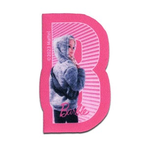 Op de afbeelding: Een roze stoffen embleem in de vorm van de letter B, met een textuur en een diagonaal gestreept patroon. Het embleem toont een afbeelding van een figuur in een grijze hoodie en de tekst Barbie en 2023 Mattel is erop gedrukt.
