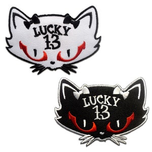 Può includere: Due toppe ricamate da stirare raffiguranti un gatto nero con occhi rossi e il testo "LUCKY 13". Una toppa è bianca con testo nero e l'altra è nera con testo bianco.
