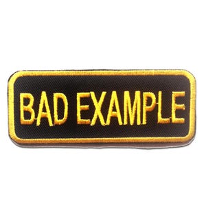 Op de afbeelding: Een rechthoekige geborduurde patch met een zwarte achtergrond en een gele rand. De tekst BAD EXAMPLE is in gele hoofdletters in het midden van de patch geborduurd. De patch heeft afgeronde hoeken en een textuur van geweven stof.