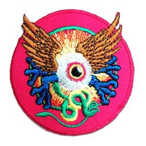 Puede incluir: Un parche circular rojo bordado con un gran ojo en el centro, rodeado por una serpiente verde y dos manos azules. El ojo tiene un iris blanco y una pupila negra. La serpiente está enroscada alrededor del ojo y tiene un cuerpo verde con detalles amarillos. Las manos están a ambos lados del ojo y tienen dedos azules. El parche está rodeado por un águila marrón con las alas extendidas.