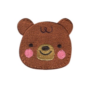 Könnte beinhalten: Brauner, gestickter Bärenaufnäher mit einem lächelnden Gesicht, goldener Nase und rosa Wangen. Der Aufnäher hat zwei kleine Ohren und ein gesticktes Design. Ideal für Kleidung oder Accessoires.