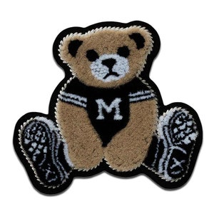 Può includere: Un orsacchiotto di peluche marrone che indossa una maglietta nera con una "M" bianca. L'orso è seduto con le gambe incrociate e indossa scarpe nere con dettagli bianchi.