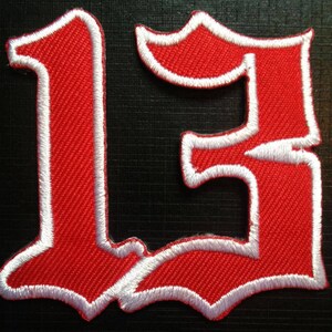 Op de afbeelding: Een rood geborduurd embleem met het nummer 13 in een gotisch lettertype. Het nummer is omrand met witte stiksels, wat een sterk contrast creëert met de rode stof. Geschikt voor kleding of accessoires.