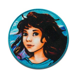 May include: A round, blue fabric patch featuring a colourful illustration of a woman with long, dark hair. The woman is looking to the right and is wearing a light blue shirt.