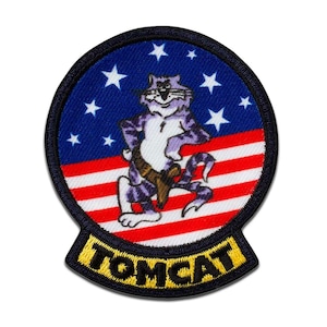 Op de afbeelding: Geborduurde patch met een paarse kat met een pistool, tegen een blauwe achtergrond met witte sterren en rood-witte strepen. Het woord "TOMCAT" wordt in gele letters op een zwarte banner weergegeven.