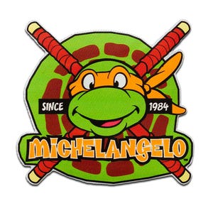 May include: A patch featuring Michelangelo from Teenage Mutant Ninja Turtles. The green turtle character wears an orange mask and is set against a green and brown background. The patch includes the text "MICHELANGELO" and "SINCE 1984". Two red and yellow nunchucks are crossed behind the character.