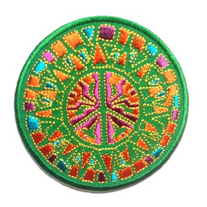 Può includere: Una toppa rotonda ricamata di colore verde con un simbolo di pace al centro. Il simbolo di pace è circondato da un design di mandala colorato con filo arancione, rosa, blu e oro.